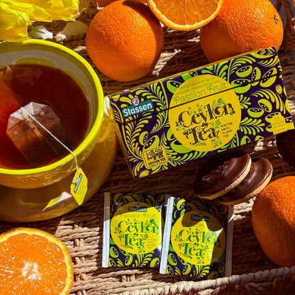 Herbata Stassen Passion Fruit Ceylon Tea w filiżance, z saszetkami, pomarańczami i opakowaniem na plecionej macie.