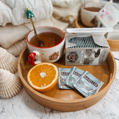 Herbata Basilur Winter Tea w saszetkach na drewnianej tacy z kubkiem naparu, pomarańczą i zimową dekoracją