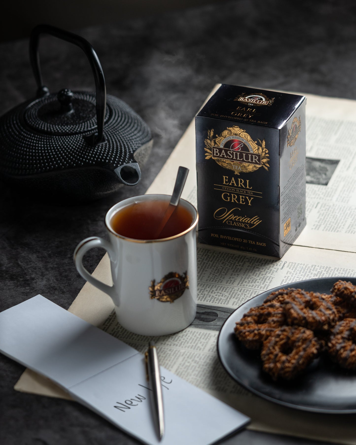 Herbata Basilur Earl Grey na gazecie obok filiżanki z gorącym naparem, żeliwnego czajnika i talerza z ciasteczkami.