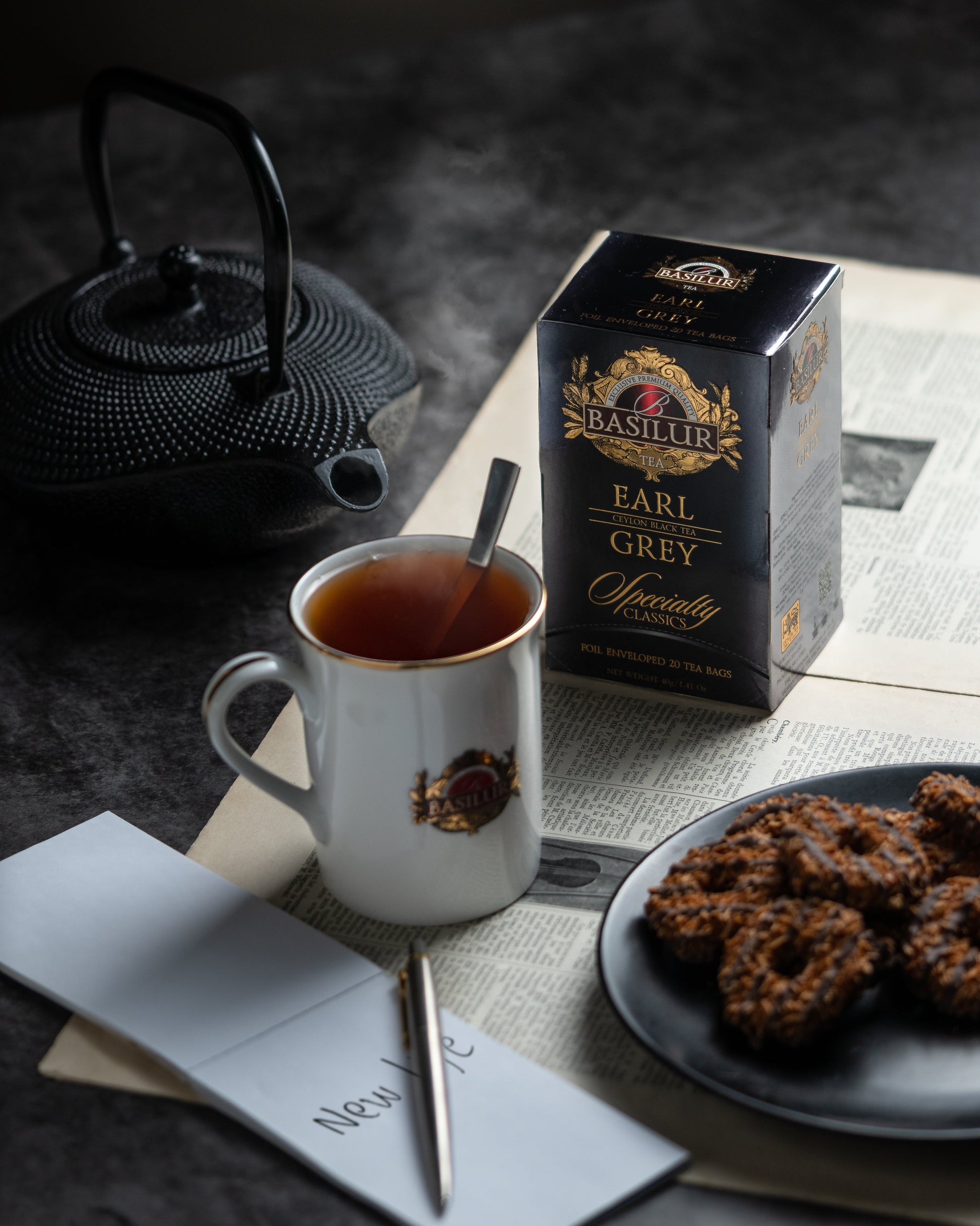 Herbata Basilur Earl Grey na gazecie obok filiżanki z gorącym naparem, żeliwnego czajnika i talerza z ciasteczkami.