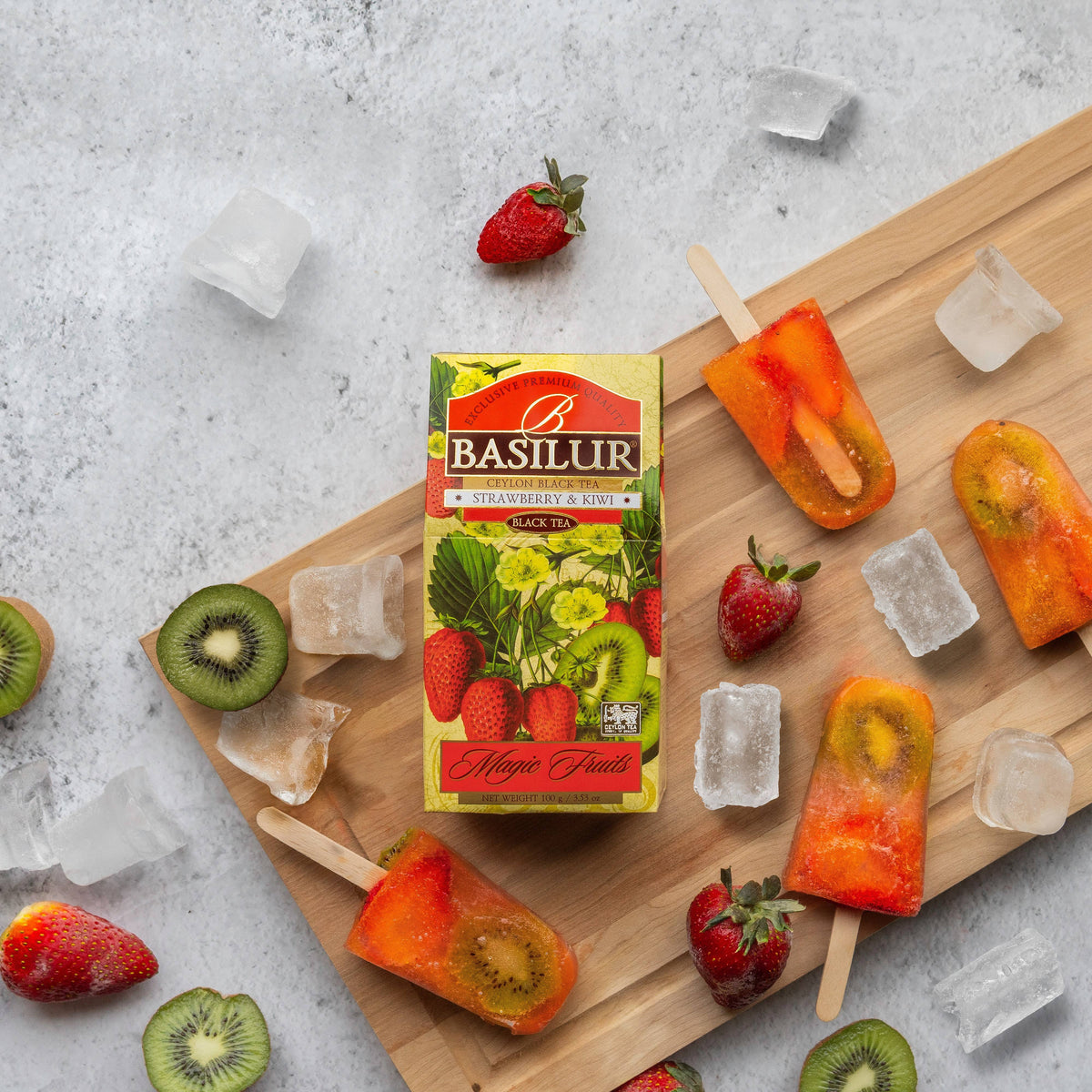 Herbata Basilur Strawberry and Kiwi na desce obok lodów z owocami, truskawek, kiwi i kostek lodu.