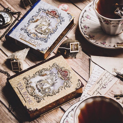 Puszka Basilur Tea Book Vol II w formie książki na drewnianym stole obok filiżanki herbaty i koperty.