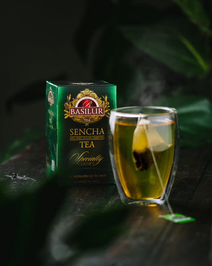 Zielone opakowanie herbaty Basilur Sencha obok szklanki z zaparzającą się torebką zielonej herbaty.