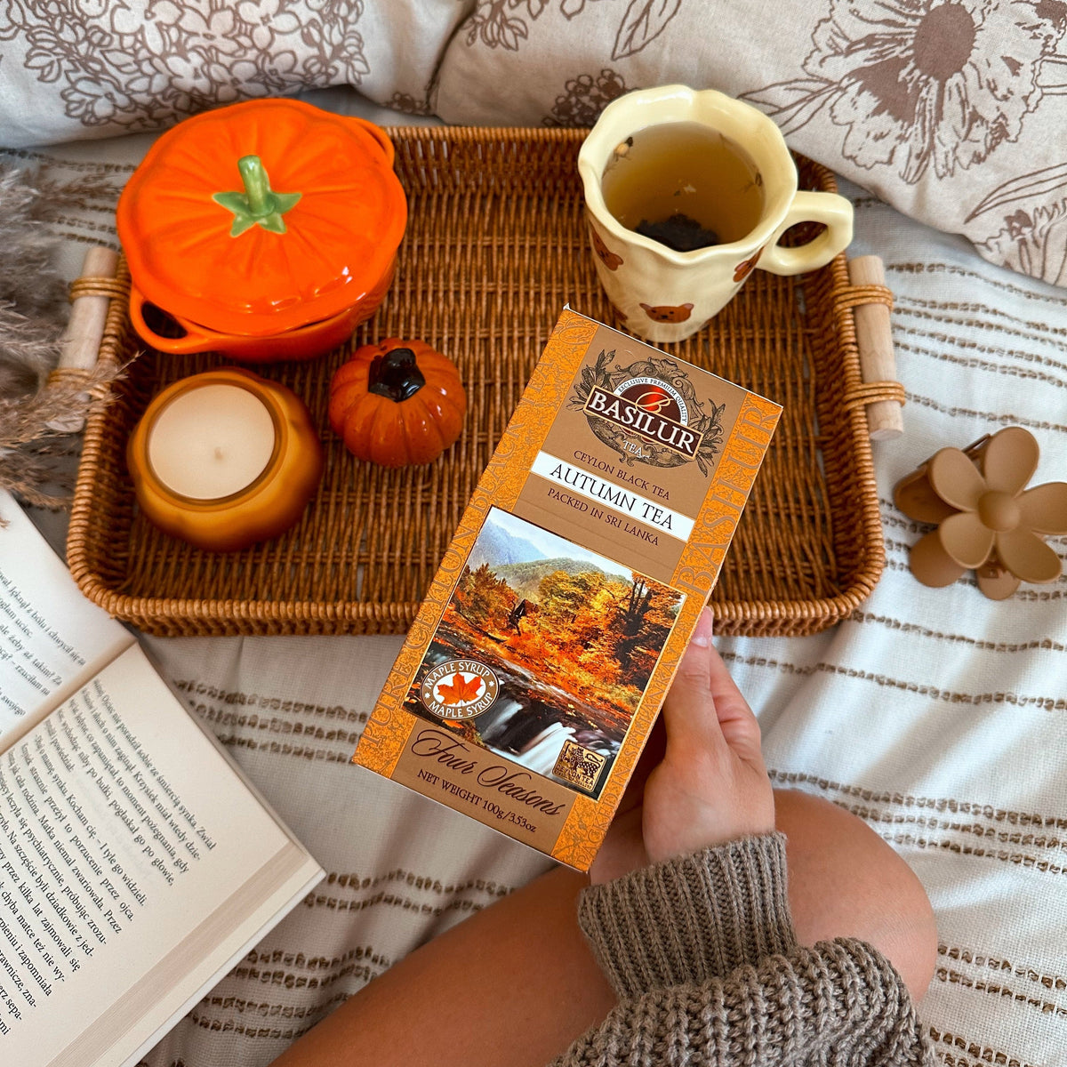 Opakowanie Basilur Autumn Tea w dłoni, na wiklinowej tacy z dyniami, świecą, książką i herbatą