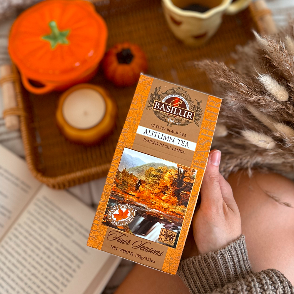 Herbata Basilur Autumn Tea w dłoni na tle jesiennych dekoracji, książki i świecy