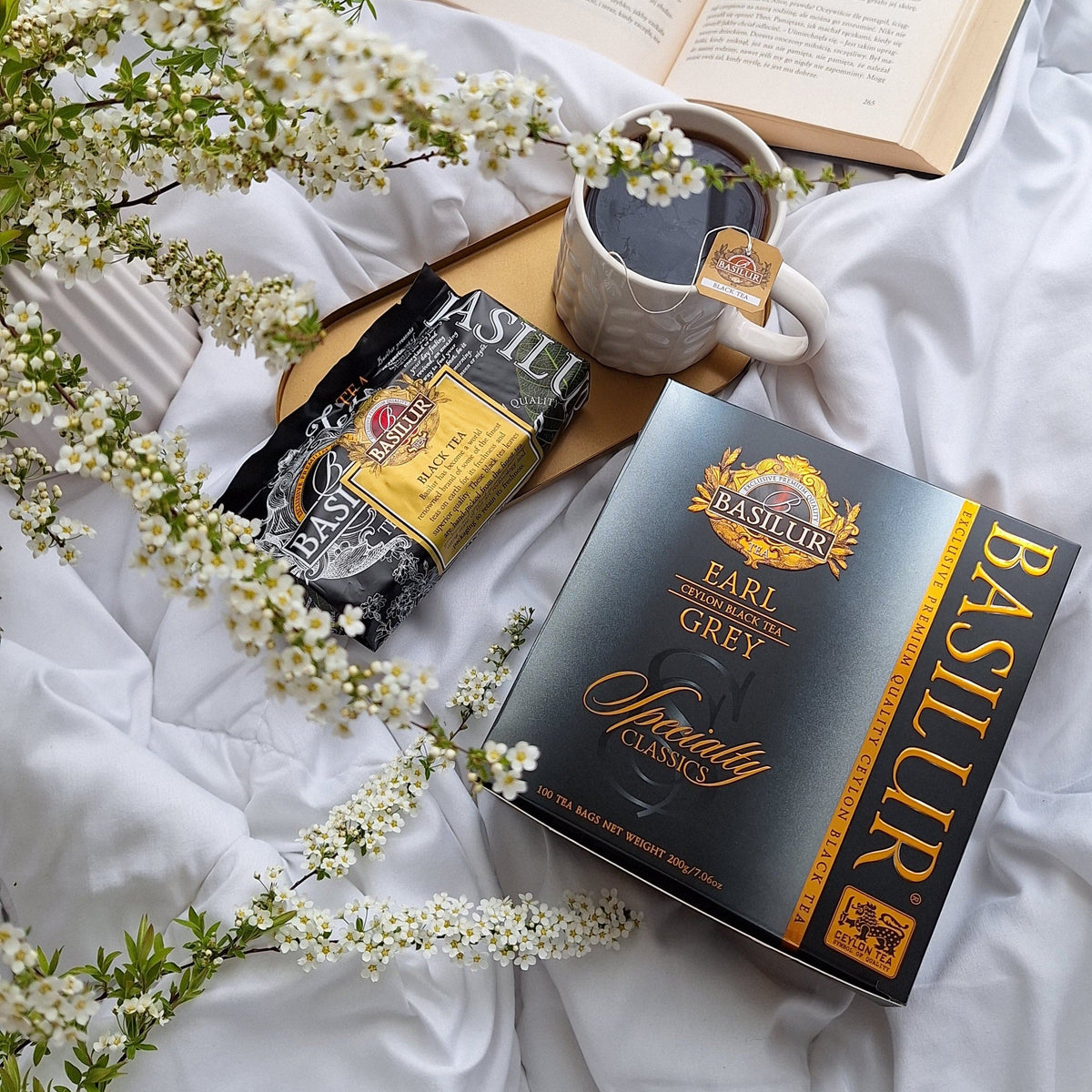 Kubek herbaty Earl Grey Basilur na białej pościeli, obok otwartej książki, opakowania herbaty i kwiatów, spokojny poranek