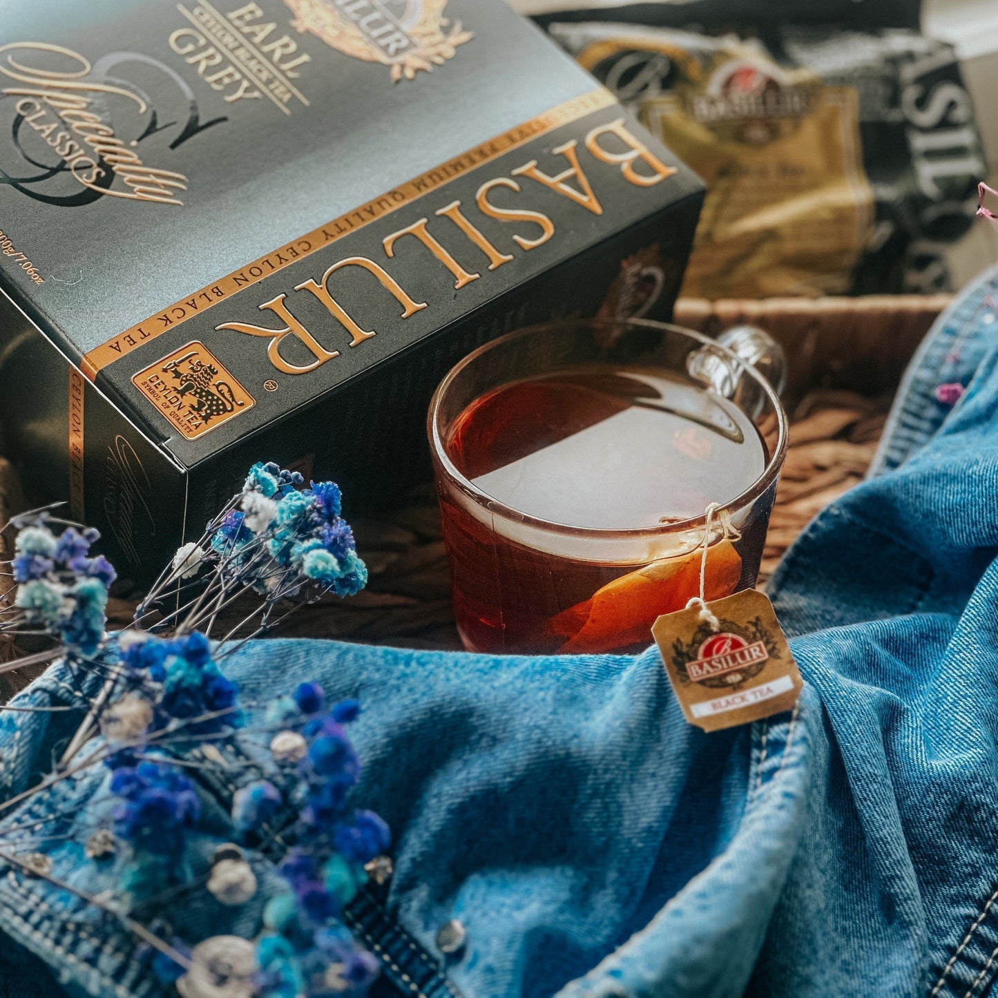Szklanka herbaty Earl Grey Basilur z cytryną obok eleganckiego opakowania, jeansu i suszonych kwiatów w klimacie lifestyle