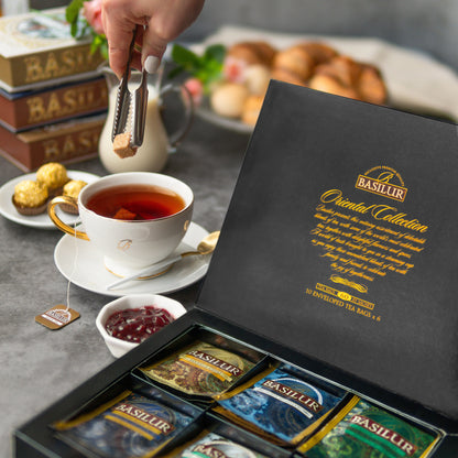 Zestaw herbat Basilur Oriental Assorted w eleganckim pudełku obok filiżanki gorącej herbaty i dodatków.
