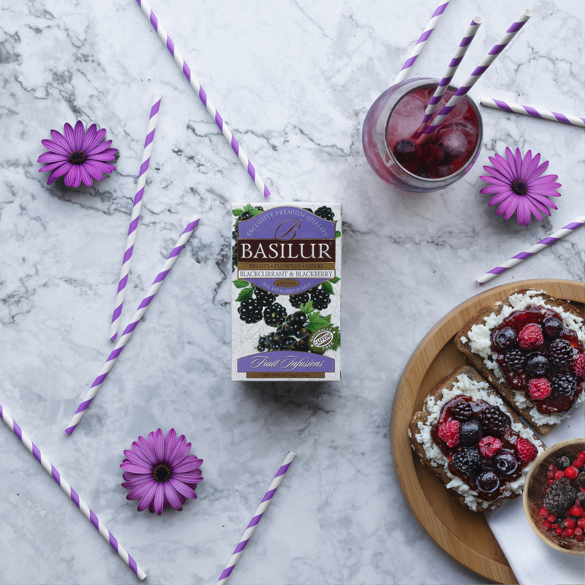 Herbata Basilur Blackcurrant and Blackberry na marmurowym tle obok kanapek z owocami, napoju i fioletowych kwiatów.