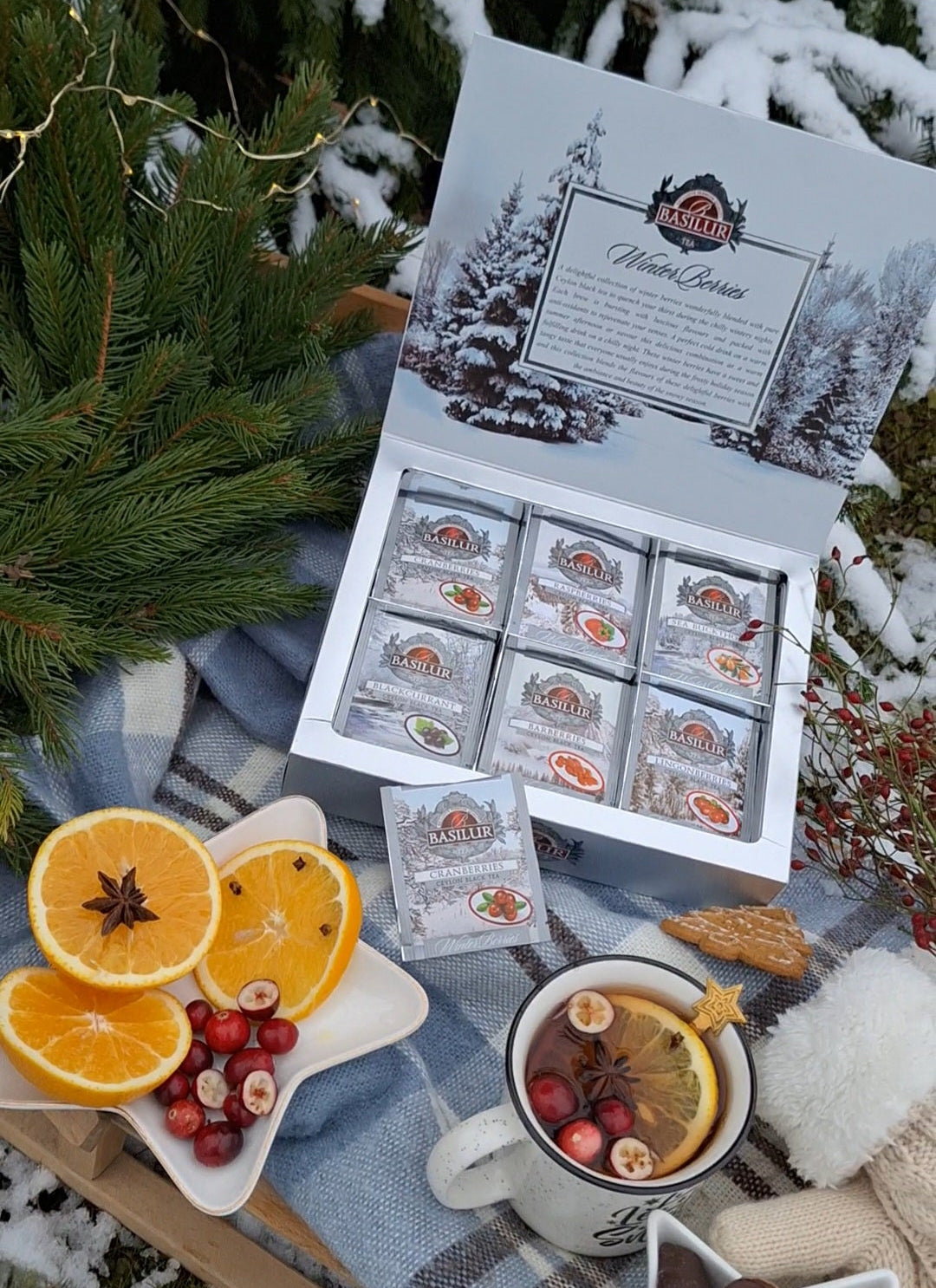 Otwarty zestaw Basilur Winter Berries na śniegu z kubkiem herbaty, pomarańczami, żurawiną i piernikami.