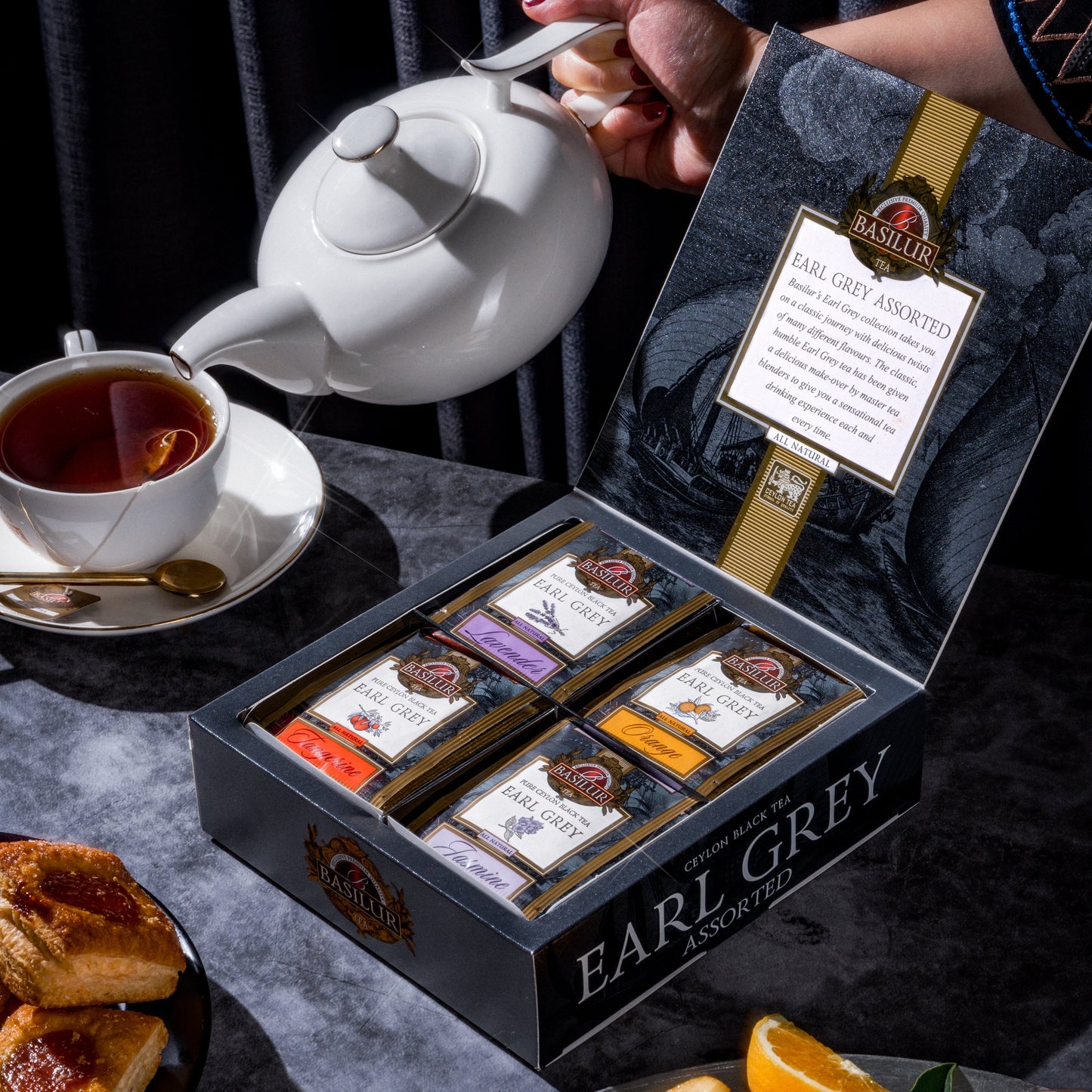 Zestaw herbat Basilur Earl Grey Assorted w otwartym pudełku obok filiżanki z gorącym naparem i owoców cytrusowych.