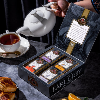 Zestaw herbat Basilur Earl Grey Assorted w otwartym pudełku obok filiżanki z gorącym naparem i owoców cytrusowych.