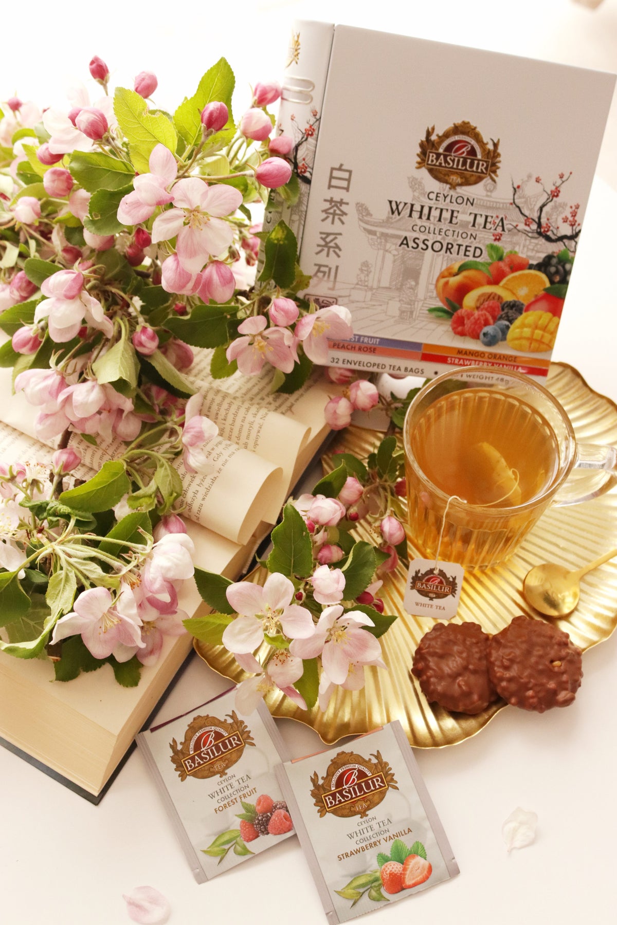 Opakowanie Basilur White Tea Assorted, filiżanka herbaty, saszetki i kwitnące gałązki na jasnym tle