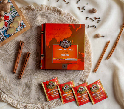 Opakowanie Basilur Breakfast Tea Asia Assorted ustawione na jasnym materialowym tle wsrod ziol i przypraw takich jak cynamon imbir gozdziki oraz galka muszkatolowa. W dolnej czesci widoczne sa czerwone saszetki herbat o roznych wariantach sniadaniowych co podkresla orientalny aromat i bogaty smak czarnej oraz bialej herbaty Basilur.